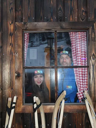 Twee personen kijken door een raam in een houten hut. Op de voorgrond zijn sleetjes zichtbaar.