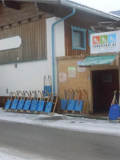 Een houten gebouw met een ingang en borden. Voor het gebouw staan veel blauwe sleeën in de sneeuw.