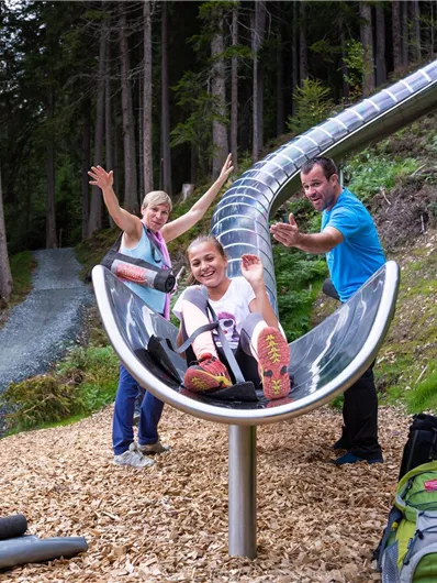 Eine Rutsche aus Metall im Wald, auf der Kinder fröhlich spielen. Im Hintergrund sind Bäume und ein Wanderweg zu sehen.