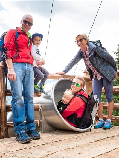 Eine Familie steht auf einer Holzplattform in den Bergen. Ein Kind sitzt in einem Rutschreifen, während die anderen lachen und die Aussicht genießen.