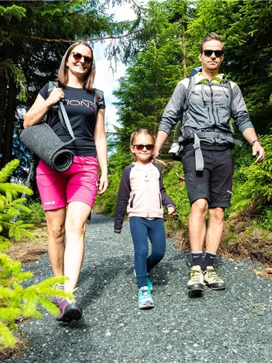 Eine Familie wandert in der Natur auf einem schmalen Weg zwischen Bäumen. Sie tragen Sonnenbrillen und entspannende Ausrüstung.