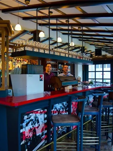 A cozy bar area with wooden shelves and stools. Two people are standing behind the counter and smiling.