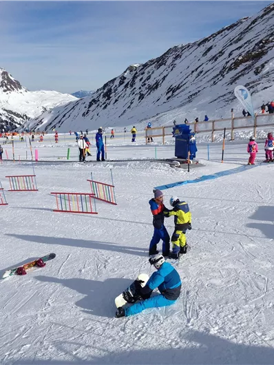 Eine verschneite Berglandschaft mit vielen Skifahrern und snowboardenden Kindern. Im Hintergrund sind hohe Berge und ein klarer Himmel zu sehen.