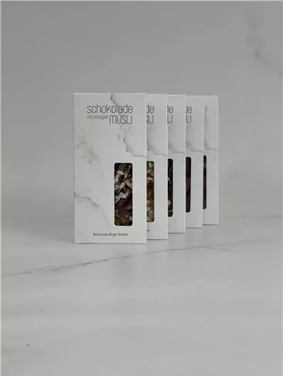 Zes verpakkingen van chocolade-muesli staan op een heldere, gladde oppervlakte. De verpakkingen hebben een elegant ontwerp met duidelijke teksten.