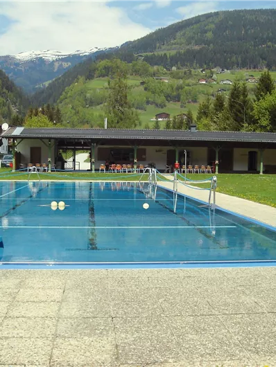 Ein Schwimmbad mit blauer Rutsche und klarem Wasser. Im Hintergrund sind grüne Hügel und Berge zu sehen.