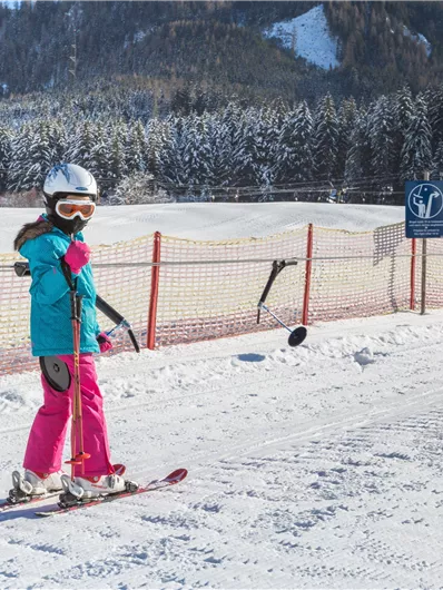 Ein Kind fährt Ski auf einer verschneiten Piste. Im Hintergrund sind Bäume und Berge sichtbar.