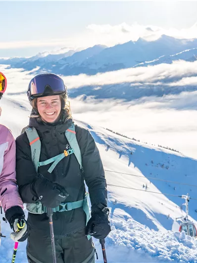 Zwei Skifahrerinnen stehen auf einem verschneiten Berg mit Blick auf die umliegenden Berge und Wolken. Die Sonne scheint und die Landschaft ist klar und beeindruckend.