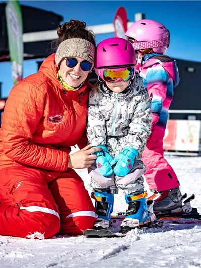 Eine lächelnde Skilehrerin in einem roten Anzug hilft einem kleinen Mädchen beim Skifahren. Im Hintergrund stehen weitere Kinder auf einer schneebedeckten Piste.