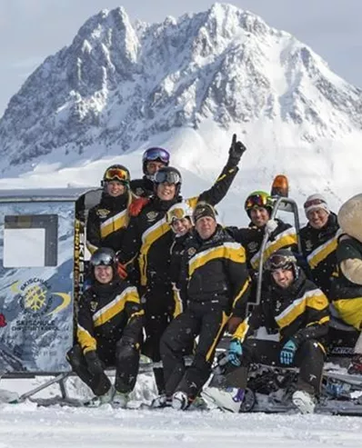 Eine Gruppe von Menschen in winterlicher Kleidung posiert vor einem Schneemobil in einer schneebedeckten Landschaft. Im Hintergrund sieht man majestätische Berge.