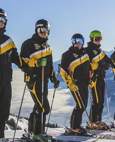 Eine Gruppe von fünf Skifahrern steht auf einem schneebedeckten Berg. Sie tragen schwarze Skianzüge mit gelben Streifen und genießen die Aussicht auf die Berge.