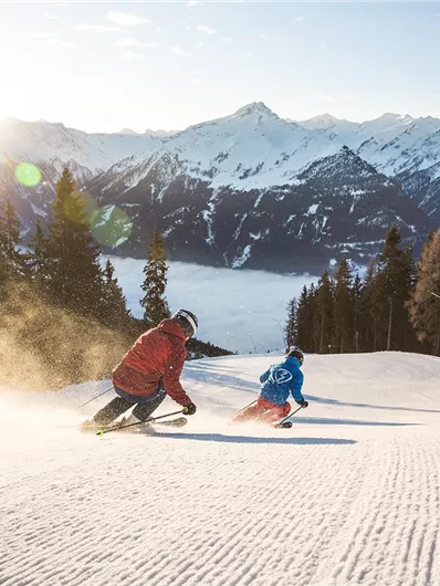 Zwei Skifahrer fahren eine Piste mit frischem Schnee hinunter. Im Hintergrund sind majestätische Berge und ein klarer Himmel zu sehen.