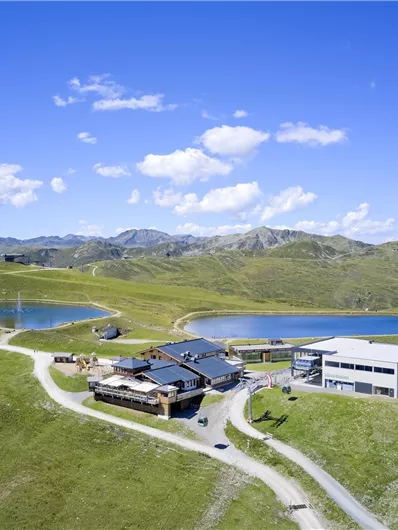 Eine malerische Berglandschaft mit sanften Hügeln und blauen Seen. Im Vordergrund sind moderne Gebäude und Wege sichtbar.