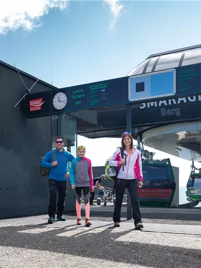 Drei Personen stehen am Eingang einer Seilbahnstation. Im Hintergrund sieht man die Seilbahn und einen klaren Himmel.