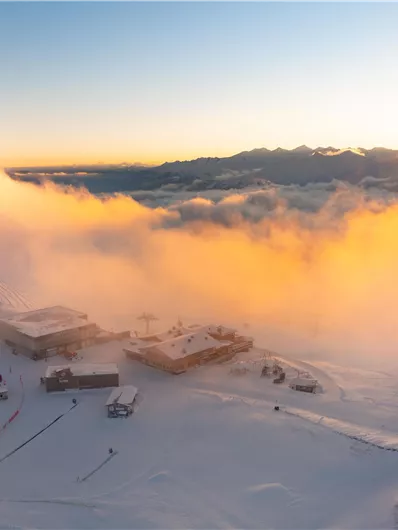 Ein verschneites Berggebiet mit Sonnenaufgang hinter den Wolken. Im Vordergrund sind mehrere Gebäude und Skilifte zu sehen.