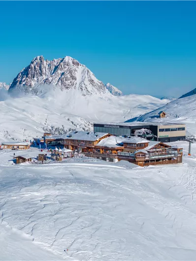 Eine verschneite Landschaft mit einem modernen Skigebäude. Im Hintergrund sind majestätische Berge unter klarem blauen Himmel zu sehen.