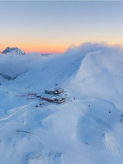 Eine verschneite Berglandschaft bei Sonnenaufgang. Im Vordergrund ist eine Skihütte zu sehen, umgeben von schneebedeckten Pisten.