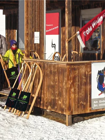 Een houten kiosk met een slee verhuur in de bergen. Familielleden staan in de sneeuw met sleetjes in de hand.