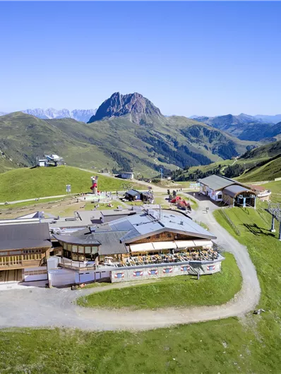 Een pittoresk berglandschap met een berghut en omringende weilanden. Op de achtergrond rijzen majestueuze bergen op onder een heldere blauwe lucht.
