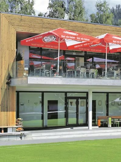 Een modern houten gebouw met een groot terras en rode luifels. De omgeving is groen en biedt een prachtig uitzicht.