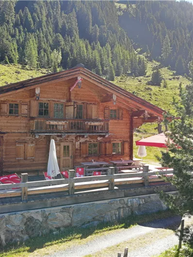 Ein rustikales Holzhaus in den Bergen umgeben von grüner Natur. Auf der Terrasse stehen einige rote Stühle und ein Sonnenschirm.