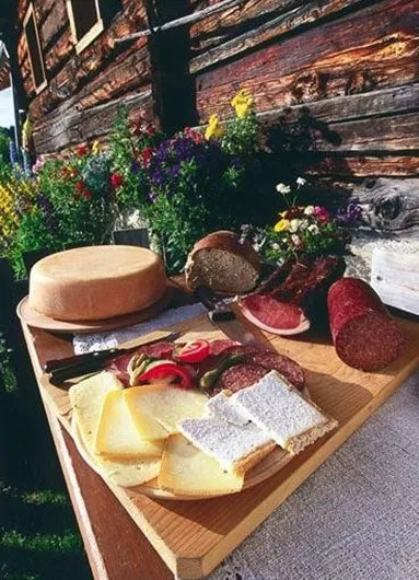 Een rustieke tafel met verschillende kazen, vleeswaren en vers brood. Op de achtergrond zijn kleurrijke bloemen en een houten schuur zichtbaar.