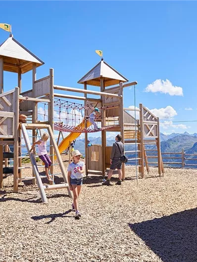 Ein Spielplatz mit Holzgerüsten und Rutschen in einem sonnigen Bergpanorama. Kinder spielen fröhlich auf dem Abenteuerplatz.