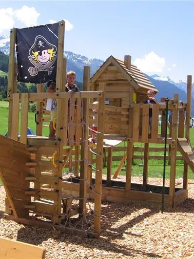 A playground with a wooden pirate castle and a slide. In the background, green meadows and mountains can be seen.