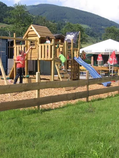 A playground with a play tower, slide, and swings in a green meadow. In the background, there is a tent with seating options.