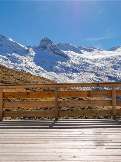 Eine malerische Berglandschaft mit schneebedeckten Gipfeln unter einem klaren blauen Himmel. Im Vordergrund ist eine Holzterrasse zu sehen.