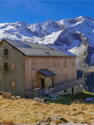 Ein rustikales Holzhaus steht in der Berglandschaft. Im Hintergrund sind schneebedeckte Berge und ein klarer blauer Himmel zu sehen.