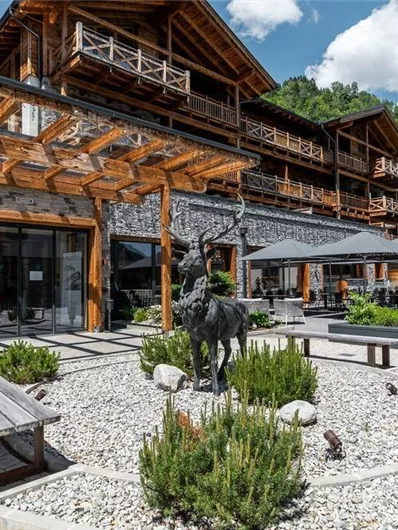 A modern lodge with a wooden facade and large windows surrounded by green spaces. In the foreground, there is a decorative garden with a stone pool and seating areas.