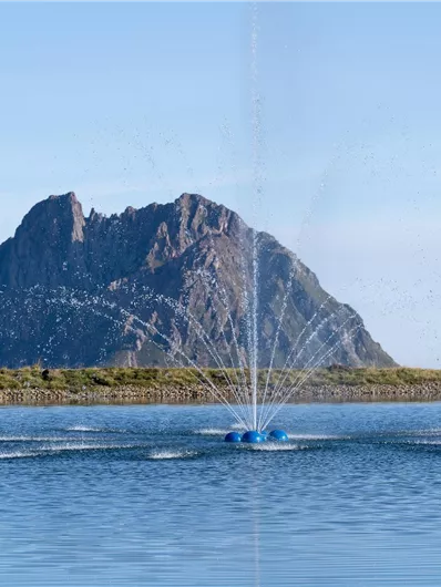 Ein ruhiger See mit einem Wasserstrahl und einer beeindruckenden Berglandschaft im Hintergrund. Wanderer genießen die Aussicht und die Natur.