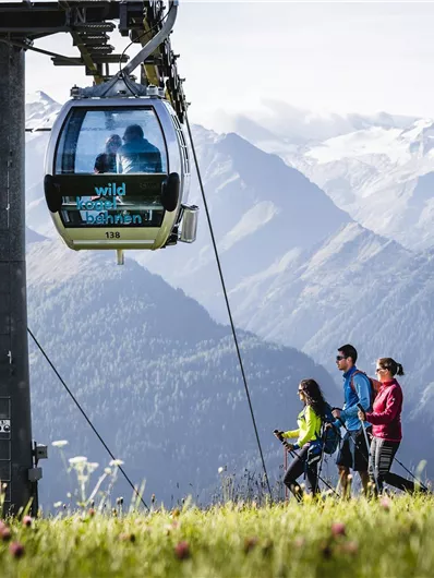 Eine Seilbahn fährt über eine grüne Wiese in den Bergen. Drei Wanderer genießen die Aussicht auf die umliegenden Berglandschaften.