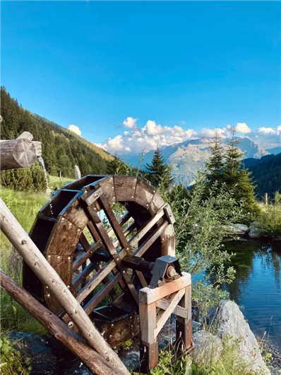 Ein Wasserrad steht am Ufer eines klaren Teichs, umgeben von grünen Wäldern und Bergen. Der Himmel ist blau mit einigen weißen Wolken.