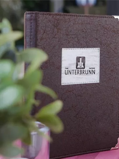 A brown, bound book titled "UNTERBRUNN". In the background, green plants and a pink tablecloth can be seen.