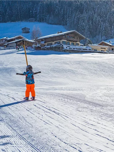 Ein verschneites Skigebiet mit Kindern, die auf der Piste spielen. Im Hintergrund sind Holzchalets und schneebedeckte Bäume zu sehen.