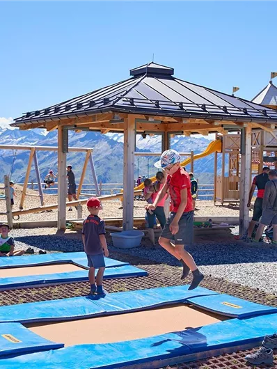 Ein Spielplatz in den Bergen mit mehreren Trampolinen. Kinder springen und spielen unter blauem Himmel und mit schöner Bergkulisse.