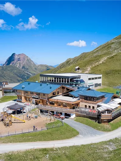 Eine alpenländische Berghütte umgeben von grünen Wiesen und Bergen. Der klare Himmel verleiht der Szenerie eine gemütliche Atmosphäre.