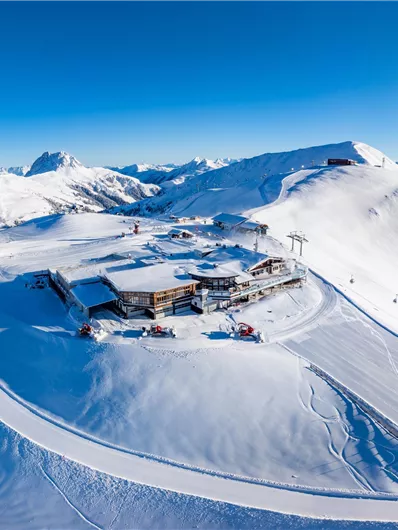Eine alpine Landschaft mit schneebedeckten Bergen und einem klaren blauen Himmel. Im Vordergrund ist ein Skigebiet mit Hütten zu sehen.
