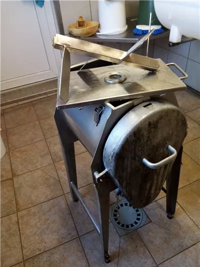 A metallic mixer stands in a kitchen on a tiled floor. It has a round container and a square lid.