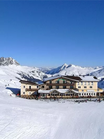 A large, elegant hotel is situated in a snow-covered landscape. In the background, there are tall mountains and a clear blue sky.