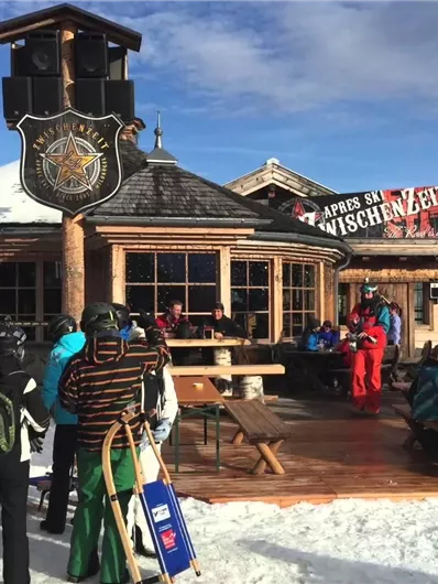 Eine rustikale Berghütte mit Sonnenterrasse in einer winterlichen Landschaft. Skifahrer und Besucher genießen die frische Luft und die Aussicht.