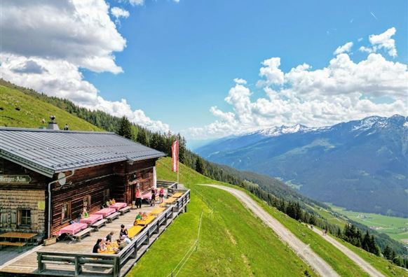 Eine gemütliche Berghütte mit Sonnenterrasse auf einer grünen Wiese. Im Hintergrund erstrecken sich die Berge unter einem blauen Himmel mit Wolken.