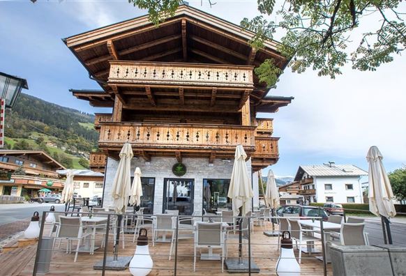 Ein charmantes Holzgebäude mit einem großen Balkon und rustikalem Stil. Vor dem Gebäude befinden sich Tische mit Stühlen und Sonnenschirmen.