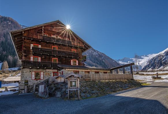 Ein traditionelles Holzhaus in den Alpen mit Blick auf schneebedeckte Berge. Die Sonne strahlt über das Dach des Hauses und der Himmel ist klar und blau.