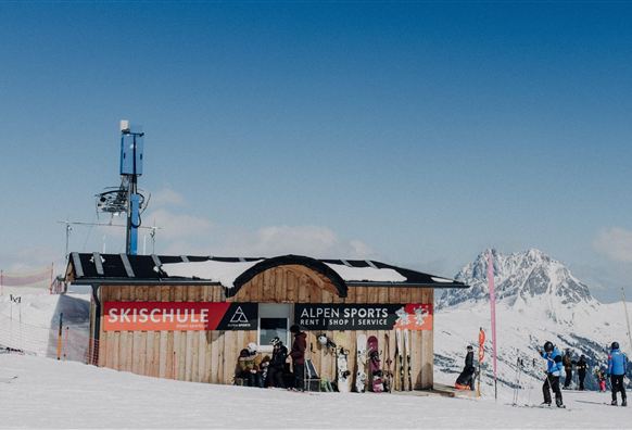 Eine Skischule mit einem Holzgebäude in den Bergen. Schnee bedeckt die Umgebung und der Himmel ist klar und blau.