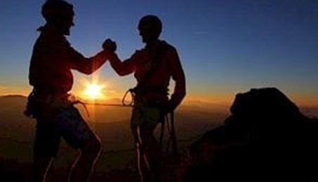 Zwei Bergsteiger feiern den Sonnenuntergang auf einem Gipfel. Sie stehen sich gegenüber und stoßen zusammen, während die Sonne im Hintergrund leuchtet.