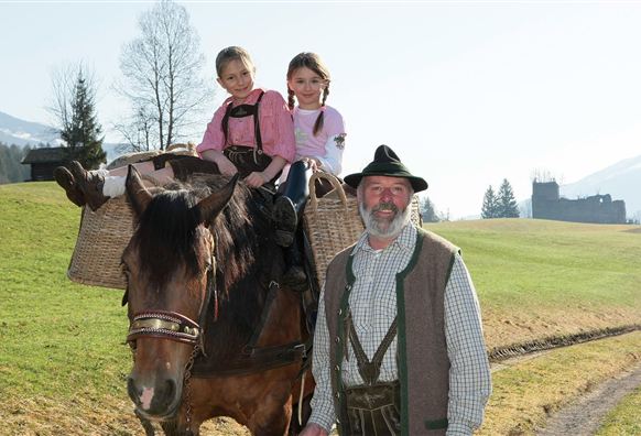 Ein Mann steht neben einem Pferd, auf dem zwei Mädchen sitzen. Die Umgebung ist ländlich mit einem klaren Himmel und grünen Wiesen.