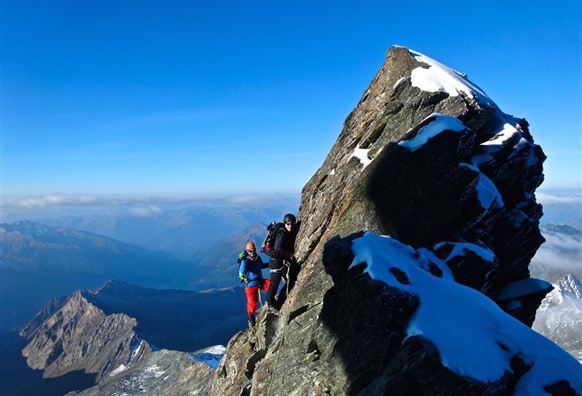 Two climbers are climbing a steep rock in the mountains. In the background, snow-covered peaks and a clear blue sky can be seen.