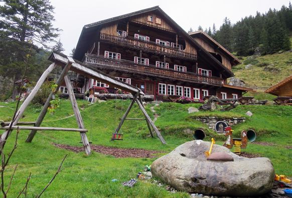 A rustic wooden house surrounded by green meadows and trees. In the foreground, there is a playground with a swing and play equipment.